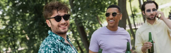 Joven con gafas de sol de pie cerca de amigos interracial con cerveza en el parque de verano, pancarta - foto de stock