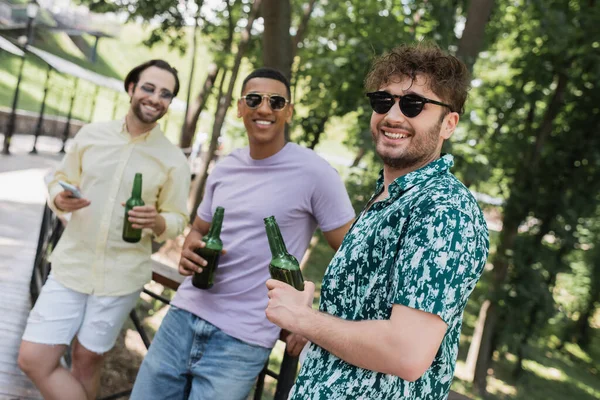 Hombre positivo sosteniendo botella de cerveza cerca de amigos interracial en gafas de sol en el parque de verano - foto de stock