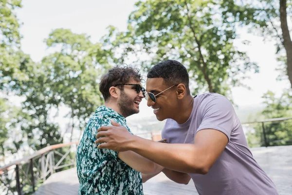 Sonriendo amigos interracial en gafas de sol abrazándose en el parque de verano - foto de stock