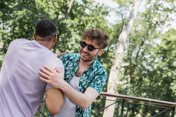 Беззаботный мужчина в солнечных очках обнимает африканского американского друга в летнем парке — стоковое фото