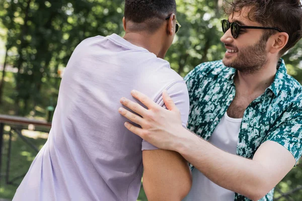Веселий молодий чоловік в сонцезахисних окулярах обіймає афроамериканського друга в літньому парку — стокове фото