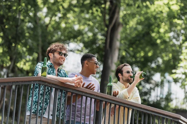 Hombre afroamericano en gafas de sol hablando con amigos cerca de la cerveza en barandilla en el parque de verano - foto de stock