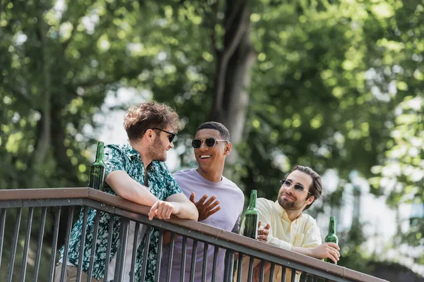 African american man in sunglasses speaking with friends near beer on railing in summer park — Stock Photo
