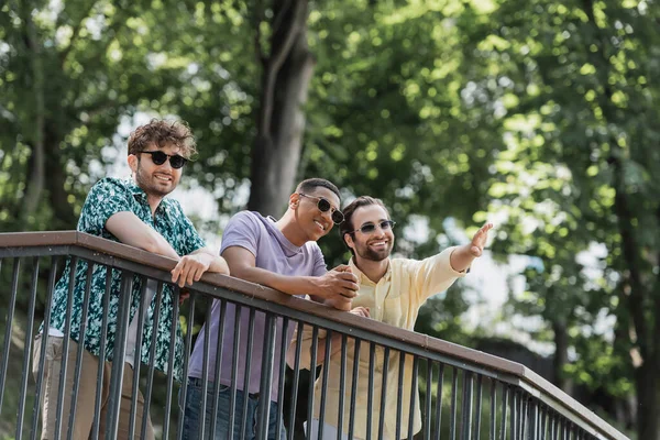 Sorrindo homem apontando com a mão perto de amigos inter-raciais em óculos de sol no parque de verão — Fotografia de Stock