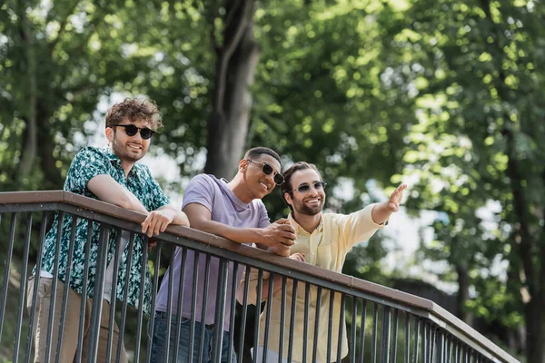 Uomo sorridente che punta con mano vicino agli amici interrazziale in occhiali da sole nel parco estivo — Foto stock