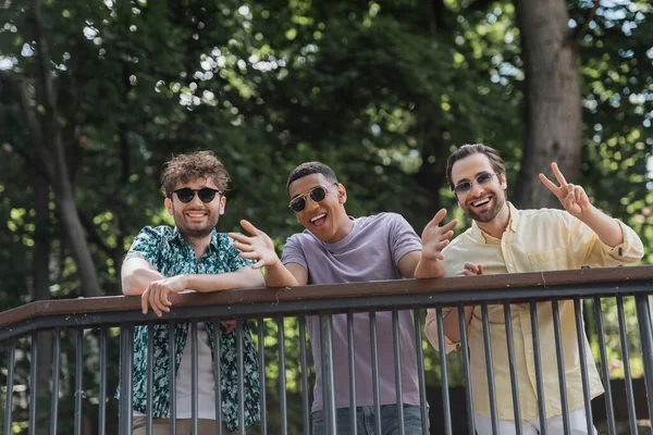 Amigos interracial positivos en gafas de sol haciendo gestos cerca de barandilla en el parque de verano - foto de stock