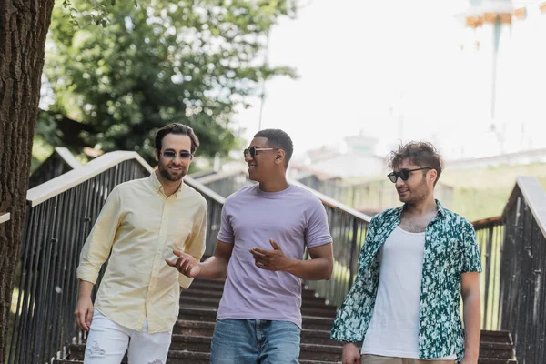 Amigos multiétnicos en gafas de sol hablando mientras caminan por las escaleras en el parque - foto de stock