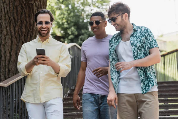 Hombre alegre usando teléfono inteligente mientras camina cerca de amigos multiétnicos en las escaleras en el parque de verano - foto de stock