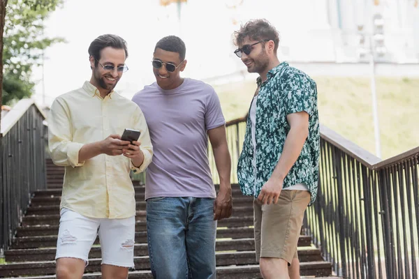 Amigos interracial despreocupados usando el teléfono celular en las escaleras en el parque de verano - foto de stock