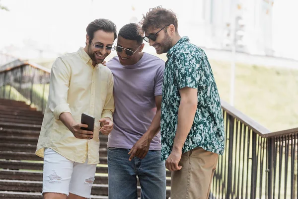 Joven mostrando teléfono celular a amigos multiétnicos en gafas de sol en las escaleras en el parque - foto de stock