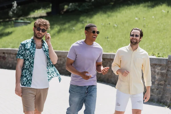 Hombres multiétnicos positivos en gafas de sol hablando mientras caminan en el parque durante el día - foto de stock