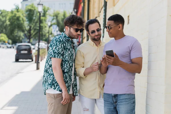 Afroamerikaner mit Sonnenbrille hält Smartphone in der Nähe von Freunden auf der Straße in Kiew — Stockfoto