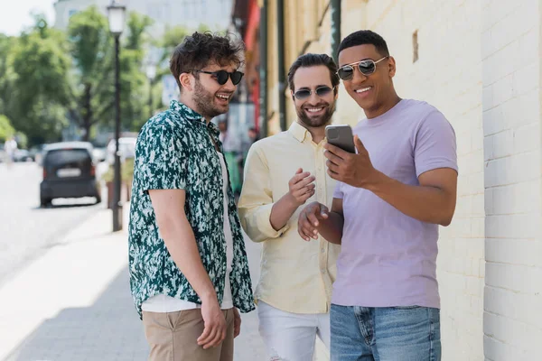 Amigos interracial despreocupados en gafas de sol con teléfono móvil en la calle en Kiev - foto de stock