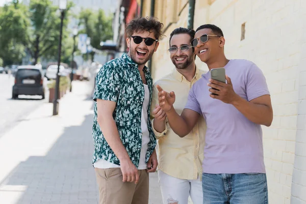 Multiethnic friends in sunglasses looking away and holding smartphone on street in Kyiv — Stock Photo
