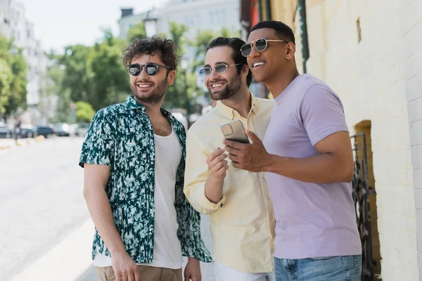Улыбающиеся межрасовые друзья в солнцезащитных очках, держащие мобильный телефон и смотрящие на улицу Киева — стоковое фото