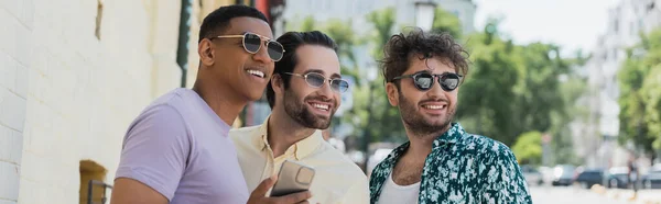 Безтурботні міжрасові друзі в сонцезахисних окулярах, використовуючи мобільний телефон і дивлячись на міську вулицю, банер — стокове фото