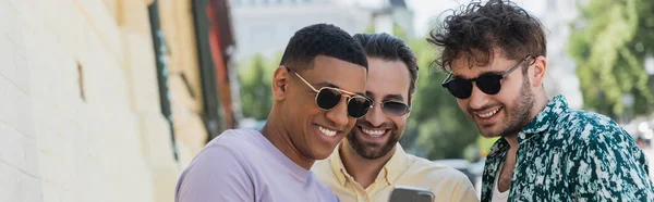 Multiethnic men in sunglasses looking at cellphone on street in Kyiv in summer, banner — Stock Photo