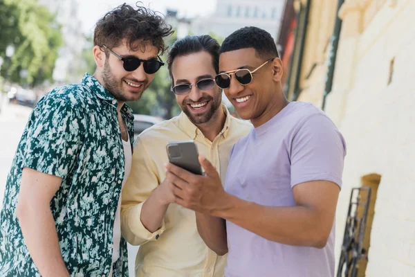 Amigos interracial positivos en gafas de sol usando teléfono móvil en la calle urbana en verano - foto de stock