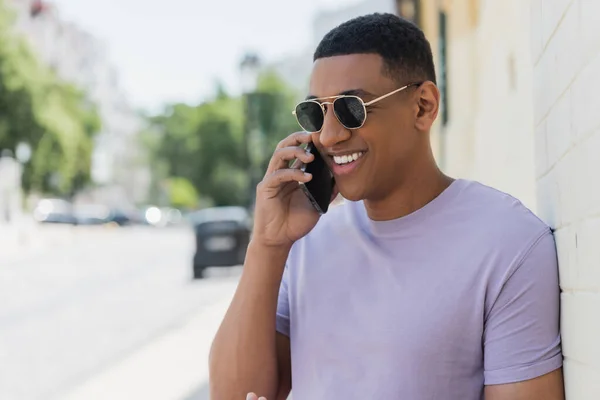 Alegre hombre afroamericano en gafas de sol hablando en el teléfono inteligente en la calle urbana borrosa - foto de stock
