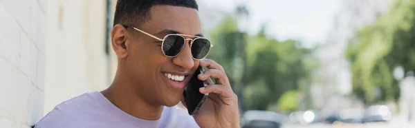 Hombre afroamericano despreocupado en gafas de sol hablando en el teléfono inteligente en la calle urbana, pancarta - foto de stock