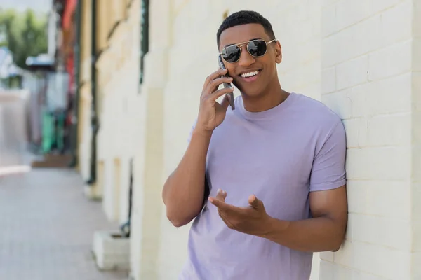 Sonriente hombre afroamericano en gafas de sol hablando por teléfono móvil en la calle urbana borrosa - foto de stock