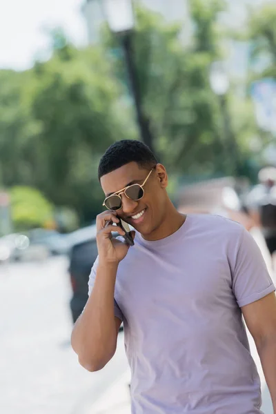 Alegre hombre afroamericano en gafas de sol hablando en teléfono inteligente en la calle urbana - foto de stock