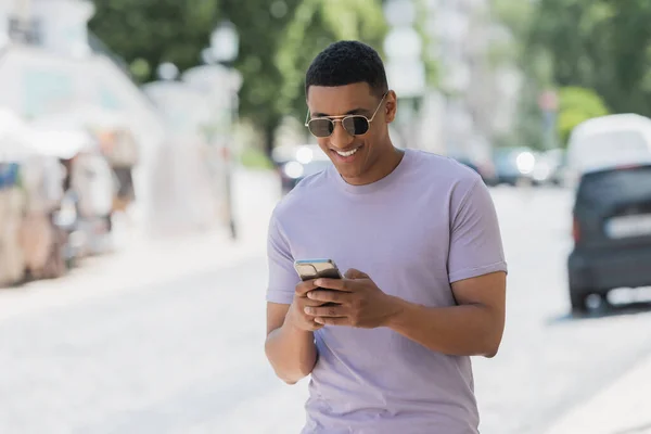 Carefree african american man in sunglasses using mobile phone on urban street — Stock Photo