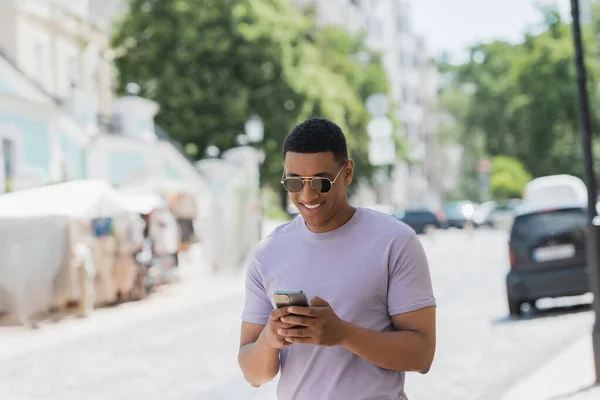 Cheerful african american man in sunglasses using smartphone while walking on urban street — Stock Photo