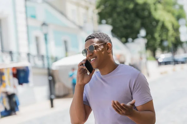 Щасливий афроамериканець у сонцезахисних окулярах говорить по смартфону на міській вулиці влітку. — стокове фото