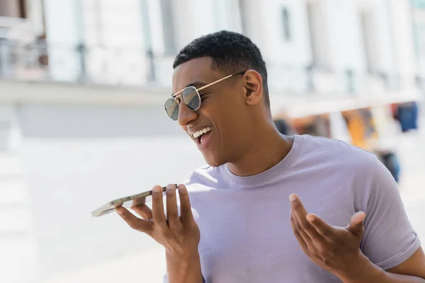 Homem americano africano alegre em óculos de sol gravando mensagem de voz no celular na rua urbana — Fotografia de Stock