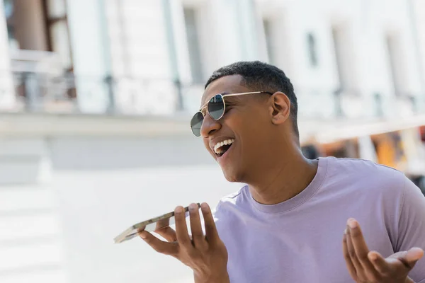 Un homme afro-américain positif avec des lunettes de soleil enregistrant un message vocal dans une rue urbaine en été — Photo de stock