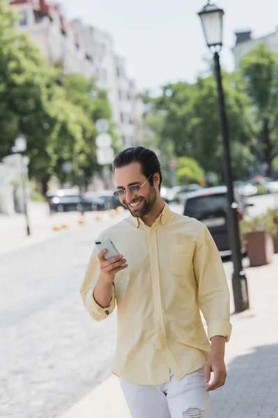 Cheerful man in shirt and sunglasses using smartphone while walking on urban street — Stock Photo