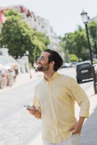Positive man in sunglasses using mobile phone while walking on blurred urban street — Stock Photo