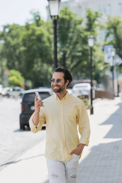 Hombre muy alegre en gafas de sol usando el teléfono celular mientras camina en la calle urbana en verano - foto de stock