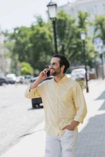 Fröhlicher junger Mann mit Sonnenbrille spricht auf verschwommener Stadtstraße mit Smartphone — Stockfoto