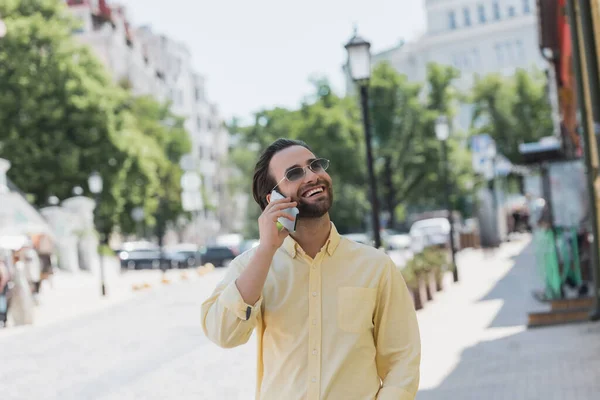 Позитивний чоловік в сонцезахисних окулярах говорить на мобільному телефоні на розмитій міській вулиці — стокове фото