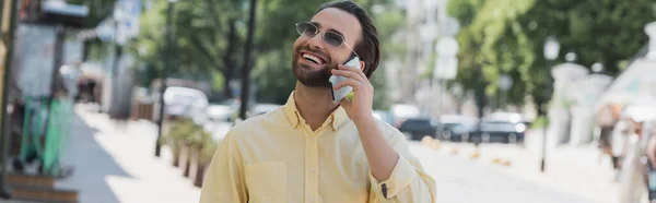Безтурботний чоловік в сонцезахисних окулярах говорить на мобільному телефоні на розмитій міській вулиці влітку, банер — стокове фото