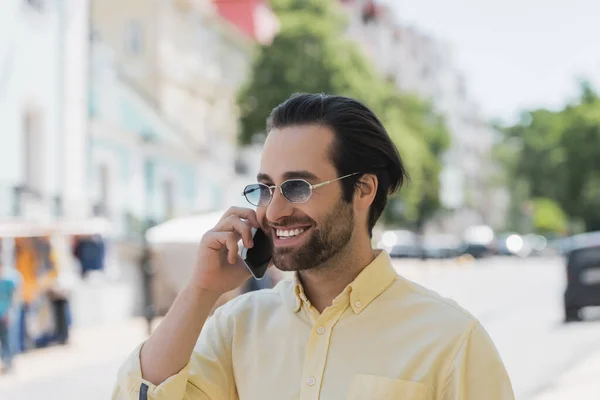 Веселий молодий чоловік в сонцезахисних окулярах говорить на смартфоні на розмитій міській вулиці — стокове фото