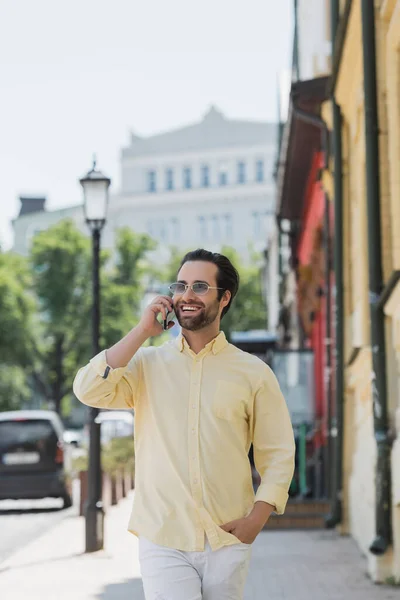 Happy brunette man in sunglasses talking on smartphone while walking on street in Kyiv — Stock Photo