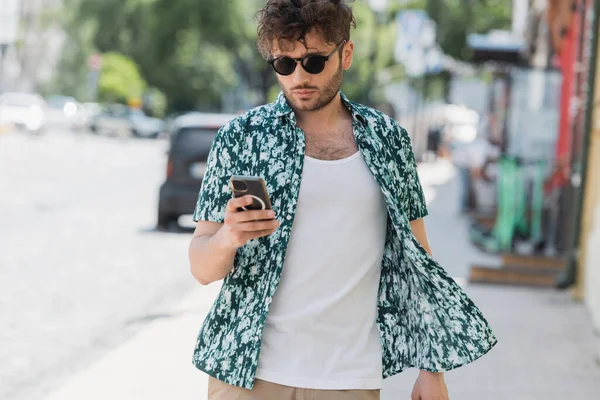Brunette man in sunglasses using smartphone while walking on urban street — Stock Photo