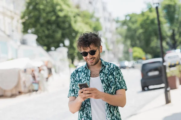 Überglücklicher Mann mit Sonnenbrille läuft und nutzt Smartphone auf Stadtstraße — Stockfoto