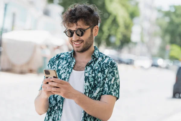 Positive brunette man in sunglasses using mobile phone on blurred urban street — Stock Photo