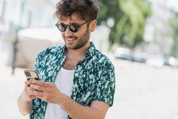 Carefree man in sunglasses using smartphone on blurred urban street in summer — Stock Photo