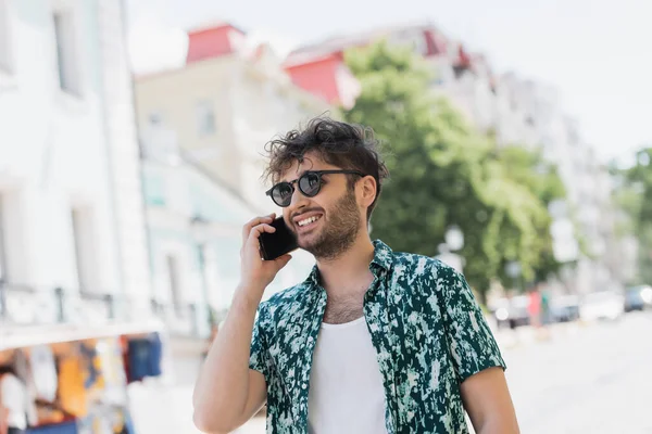 Позитивный молодой человек в солнечных очках разговаривает по смартфону во время прогулки по городской улице — стоковое фото