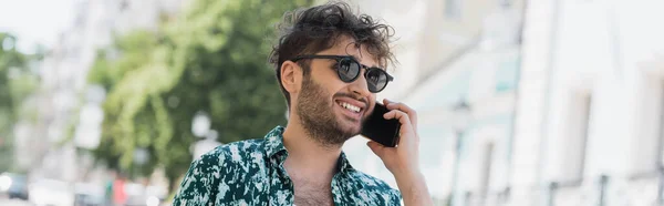 Веселый молодой человек в солнечных очках разговаривает по смартфону во время прогулки по городской улице, баннер — стоковое фото