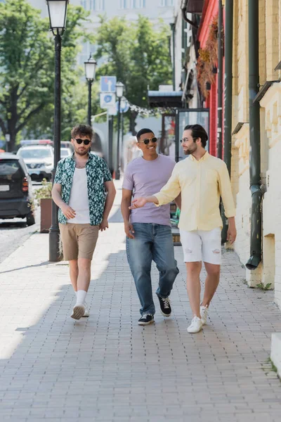 Smiling interracial friends in sunglasses walking on Andrews descent in Kyiv — Stock Photo