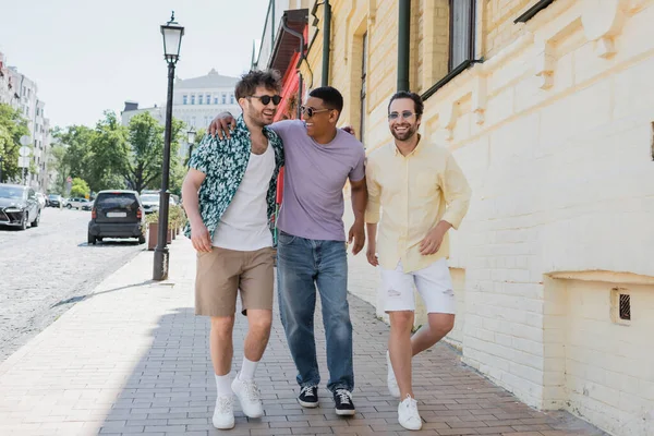Посміхнені міжрасові друзі в сонцезахисних окулярах ідуть тротуаром по Андріївському спуску в Києві. — стокове фото