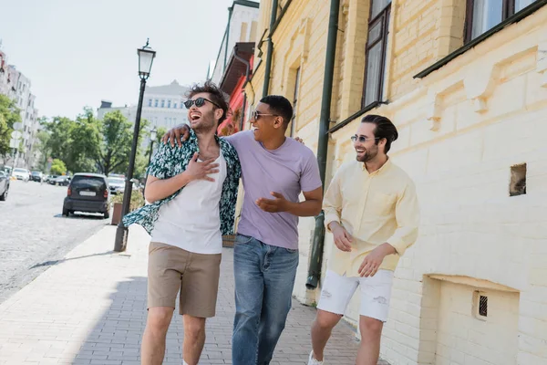 Positive multiethnische Freunde mit Sonnenbrille umarmen sich und gehen auf der Andreasabfahrt in Kiew — Stockfoto