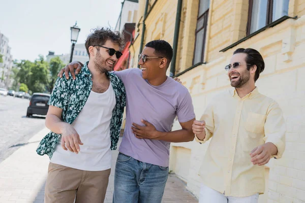 Cheerful multiethnic friends in sunglasses walking on blurred Andrews descent in Kyiv — Stock Photo