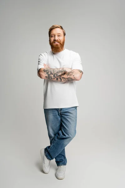 Повна довжина усміхненого татуйованого чоловіка, що перетинає руки на сірому фоні — стокове фото